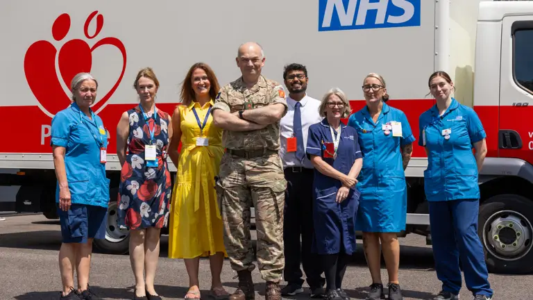 Group of NHS staff and a military personnel standing in front of a blood donation truck on a sunny day.