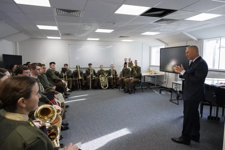 A man in a suit speaks to a group of uniformed musicians seated with brass instruments in a bright room, creating an engaged and formal atmosphere.