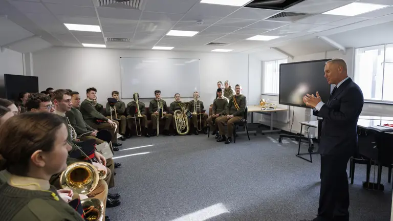 A man in a suit speaks to a group of uniformed musicians seated with brass instruments in a bright room, creating an engaged and formal atmosphere.