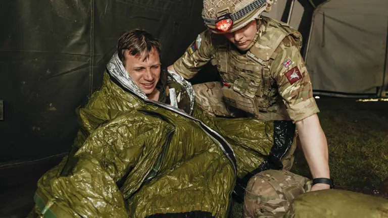 Soldier in camouflage uniform assisting a person wrapped in a green emergency thermal blanket inside a tent.