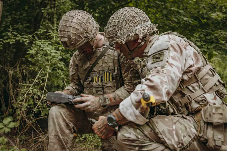 Two soldiers in camouflage gear operate a Parrot drone controller while crouched in a forested area.