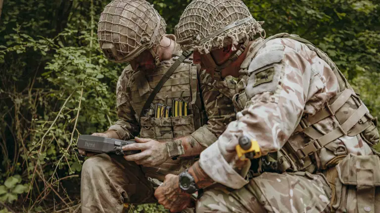 Two soldiers in camouflage gear operate a Parrot drone controller while crouched in a forested area.