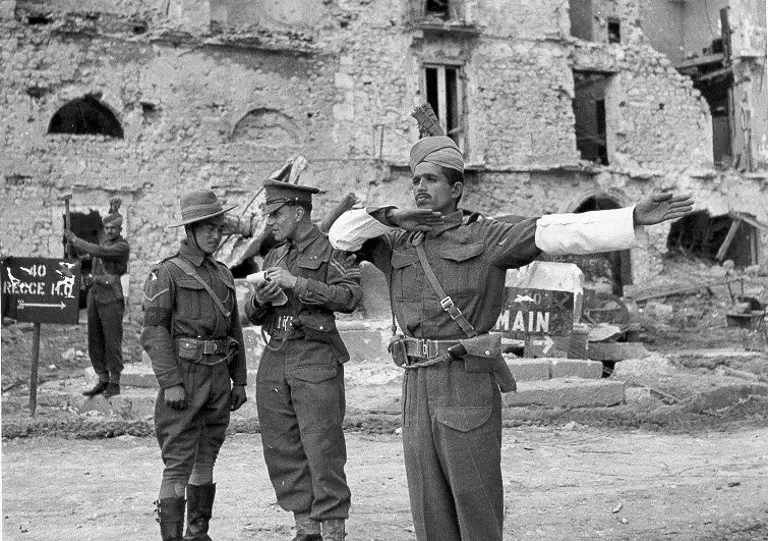 A person in a military uniform directs traffic with arms outstretched in front of a damaged building. Two others in uniform stand nearby, one reading a document and the other adjusting gear. A sign with