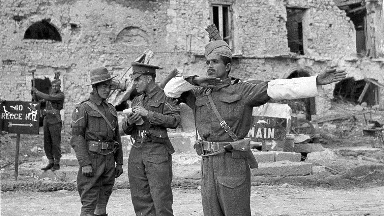 A person in a military uniform directs traffic with arms outstretched in front of a damaged building. Two others in uniform stand nearby, one reading a document and the other adjusting gear. A sign with