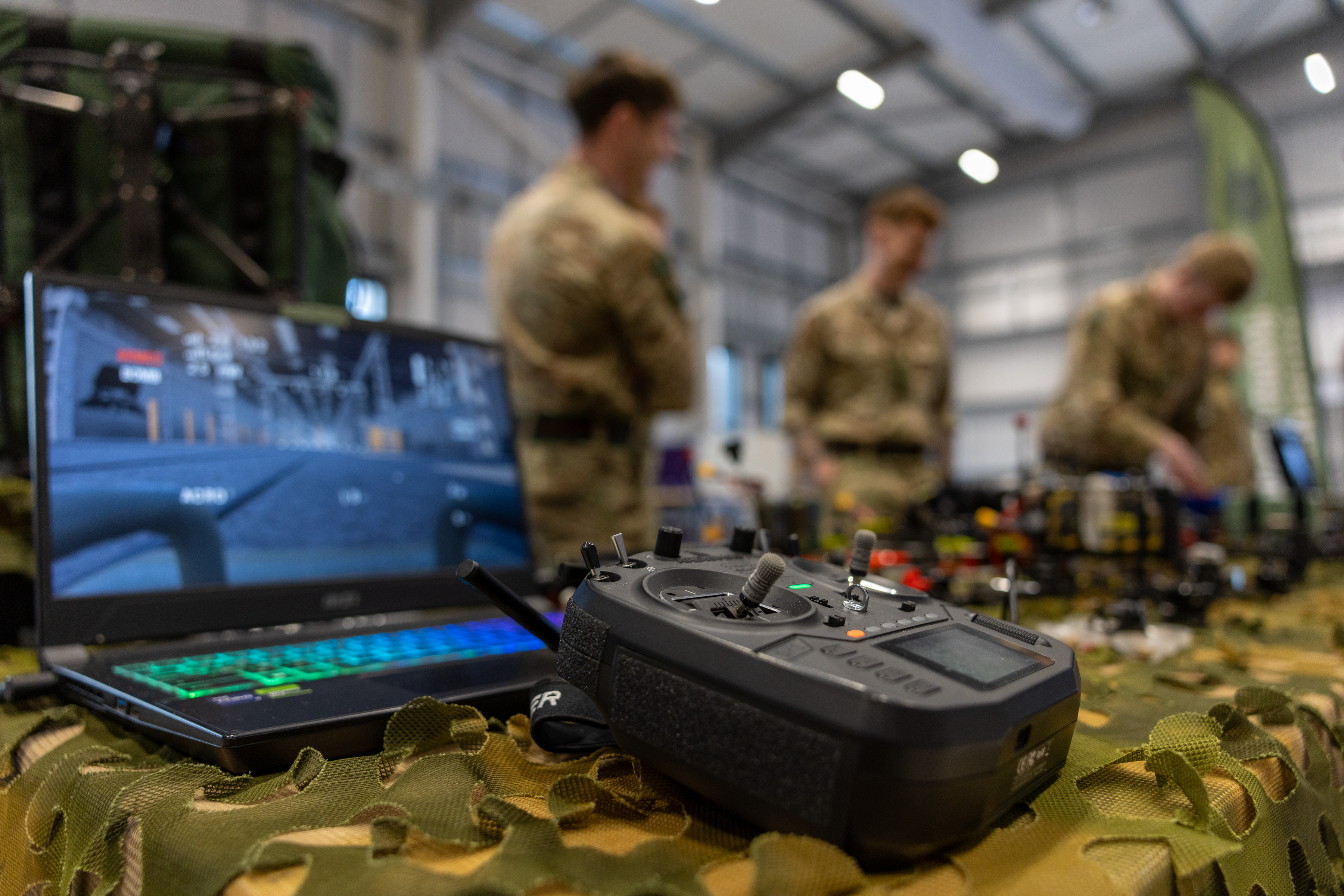 Military drone controller and laptop with simulation software on a camouflaged table inside a training facility.