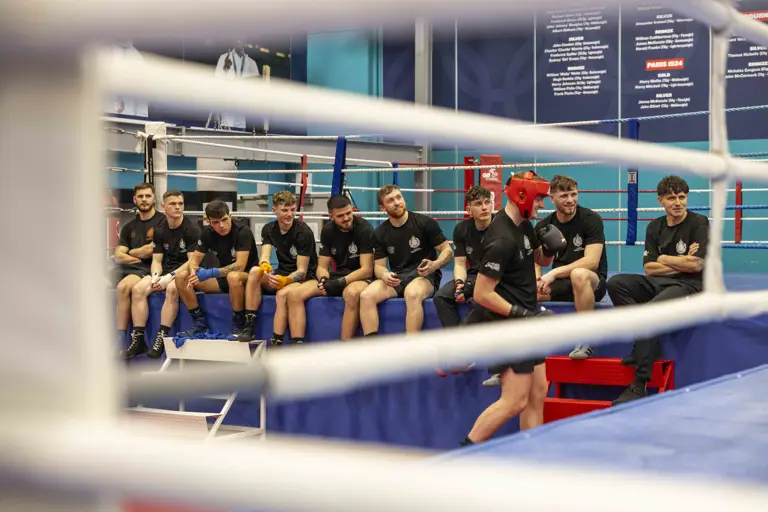 A group of men wearing black t-shirts and shorts sit on the edge of a boxing mat.