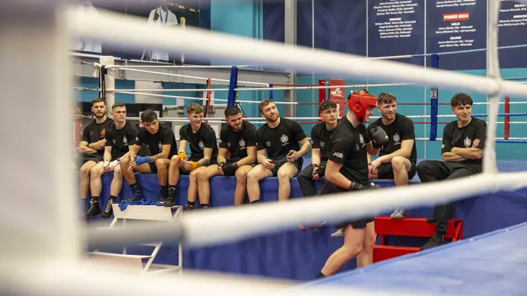 A group of men wearing black t-shirts and shorts sit on the edge of a boxing mat.