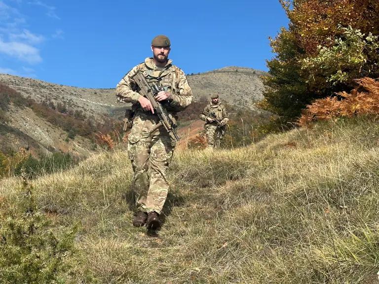 Two soldiers, wearing camouflage uniforms and carrying rifles, walk through a field.