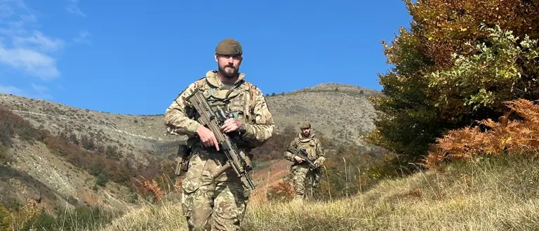 Two soldiers, wearing camouflage uniforms and carrying rifles, walk through a field.