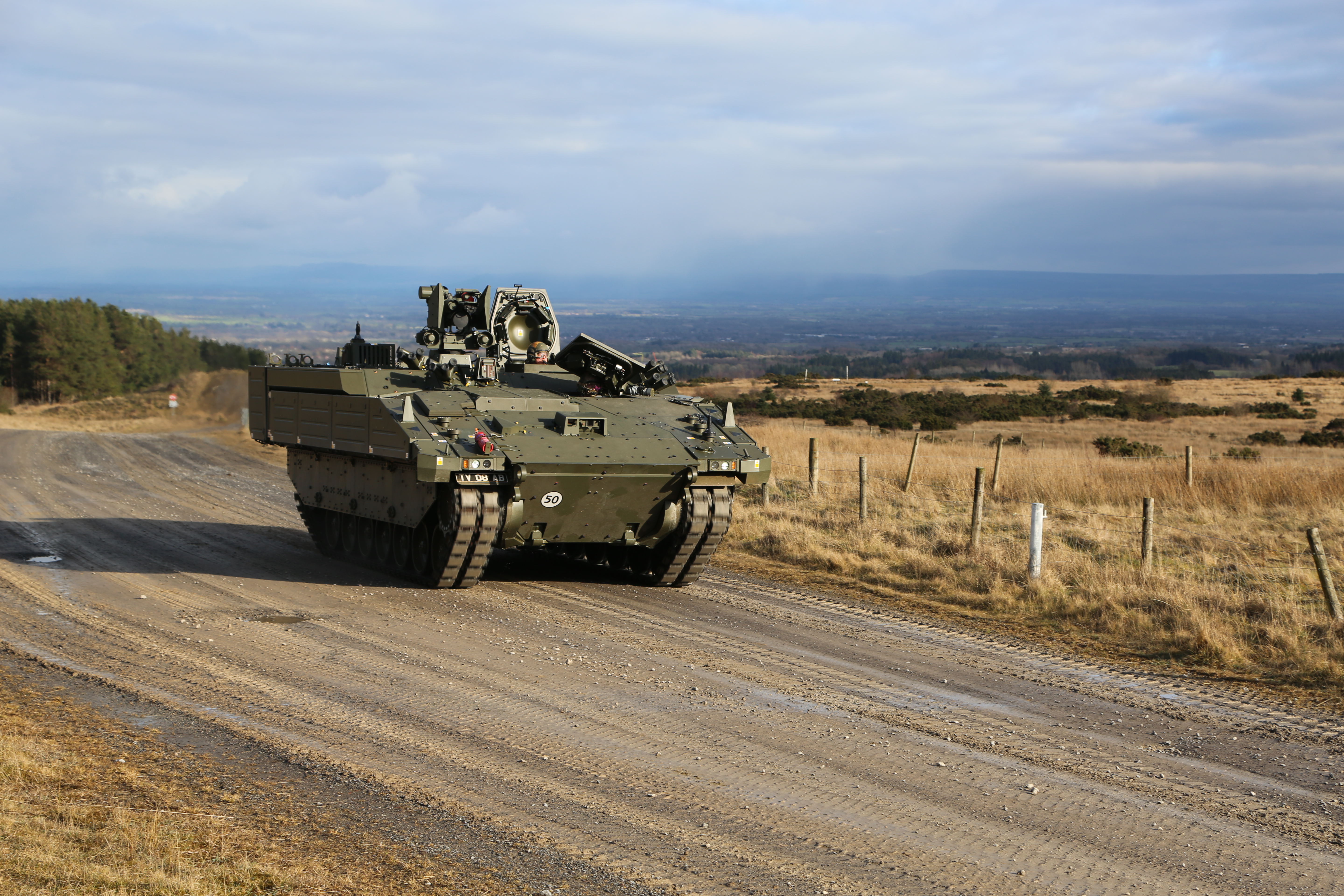 A green armoured vehicle drives along a track between two fields.