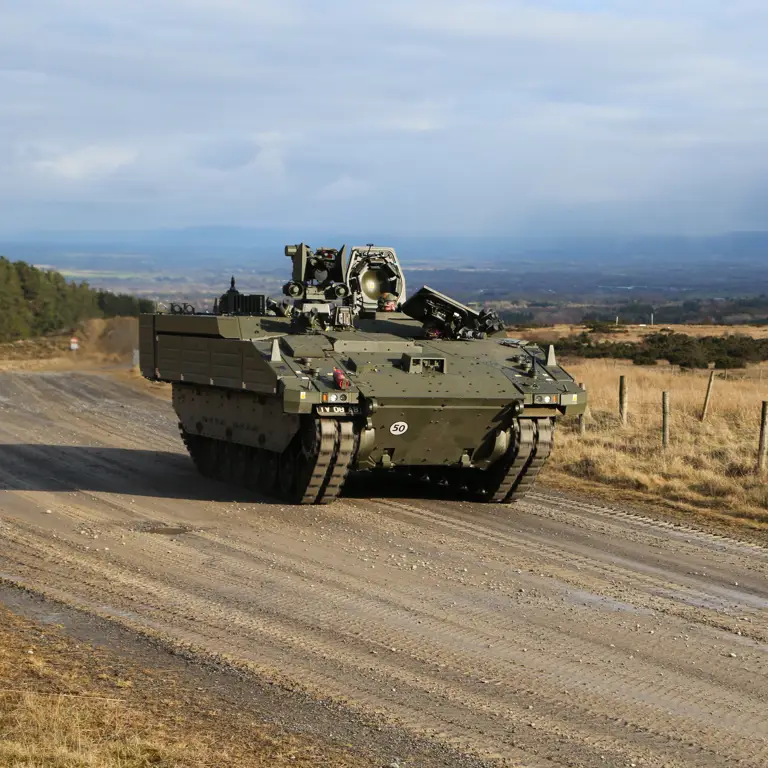 A green armoured vehicle drives along a track between two fields.