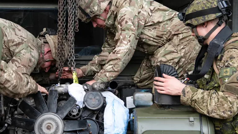 Three soldiers in camouflage uniforms and helmets work on a military vehicle engine. They appear focused and cooperative, using chains to lift parts.