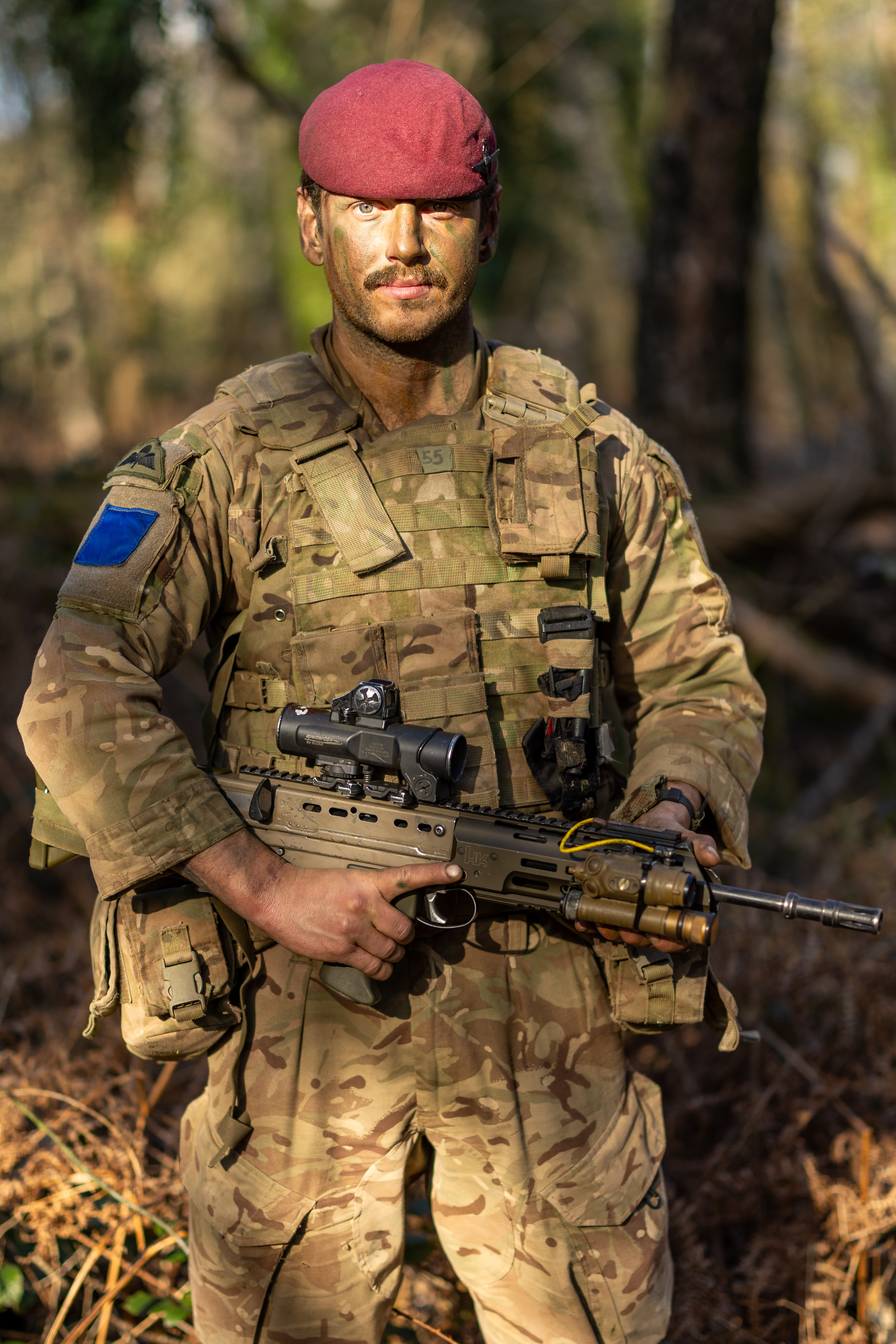 A person in military camouflage holds a rifle while standing in a forest. The individual is wearing a maroon beret.
