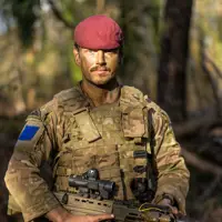 A person in military camouflage holds a rifle while standing in a forest. The individual is wearing a maroon beret.
