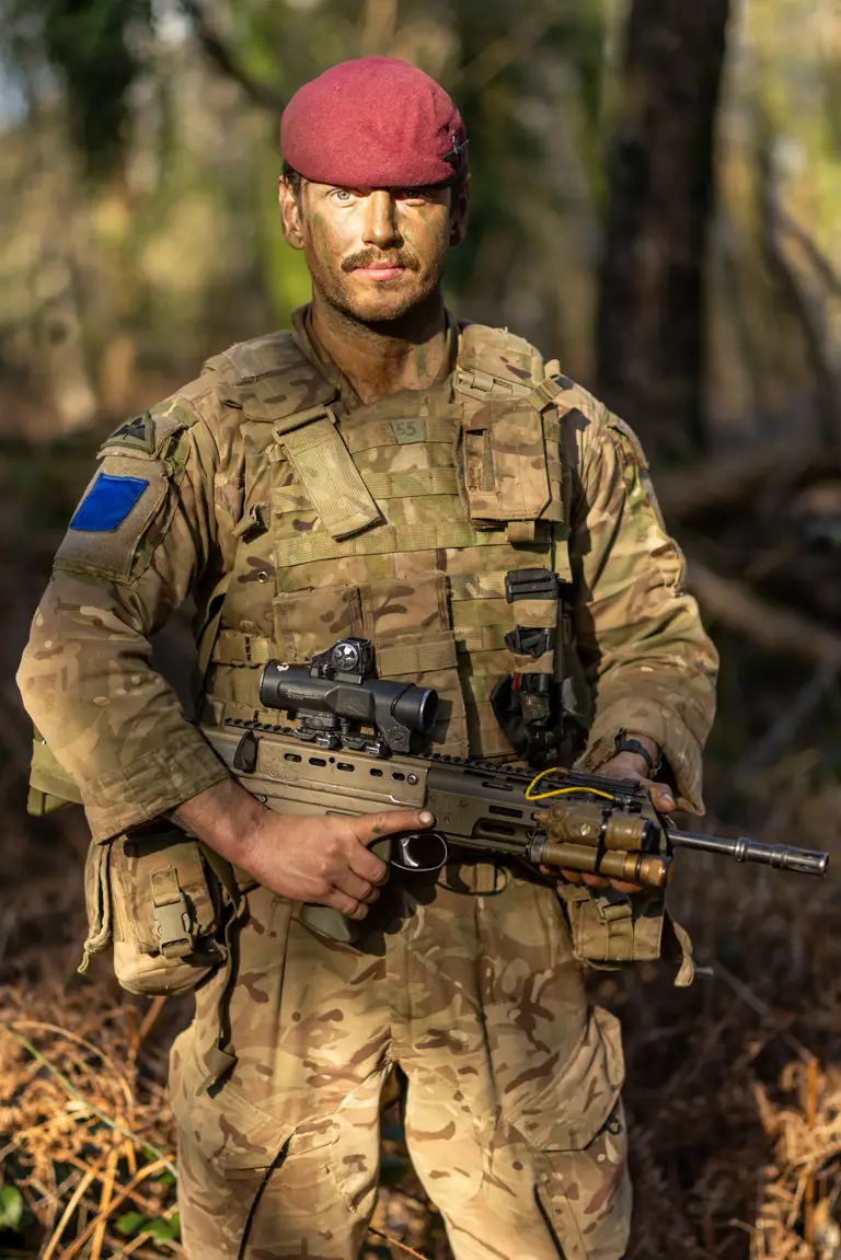 A person in military camouflage holds a rifle while standing in a forest. The individual is wearing a maroon beret.