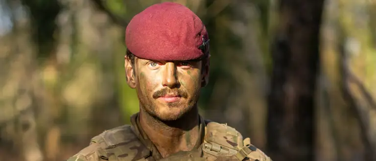 A person in military camouflage holds a rifle while standing in a forest. The individual is wearing a maroon beret.