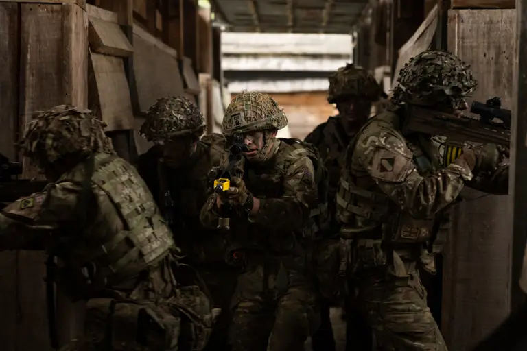 Five soldiers in camouflage uniform and helmets all facing outwards in various directions with weapons at the reason while just entering out a dark building.