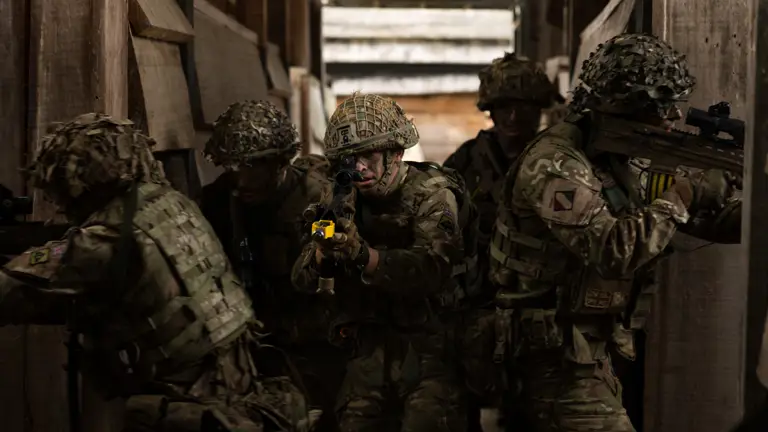 Five soldiers in camouflage uniform and helmets all facing outwards in various directions with weapons at the reason while just entering out a dark building.