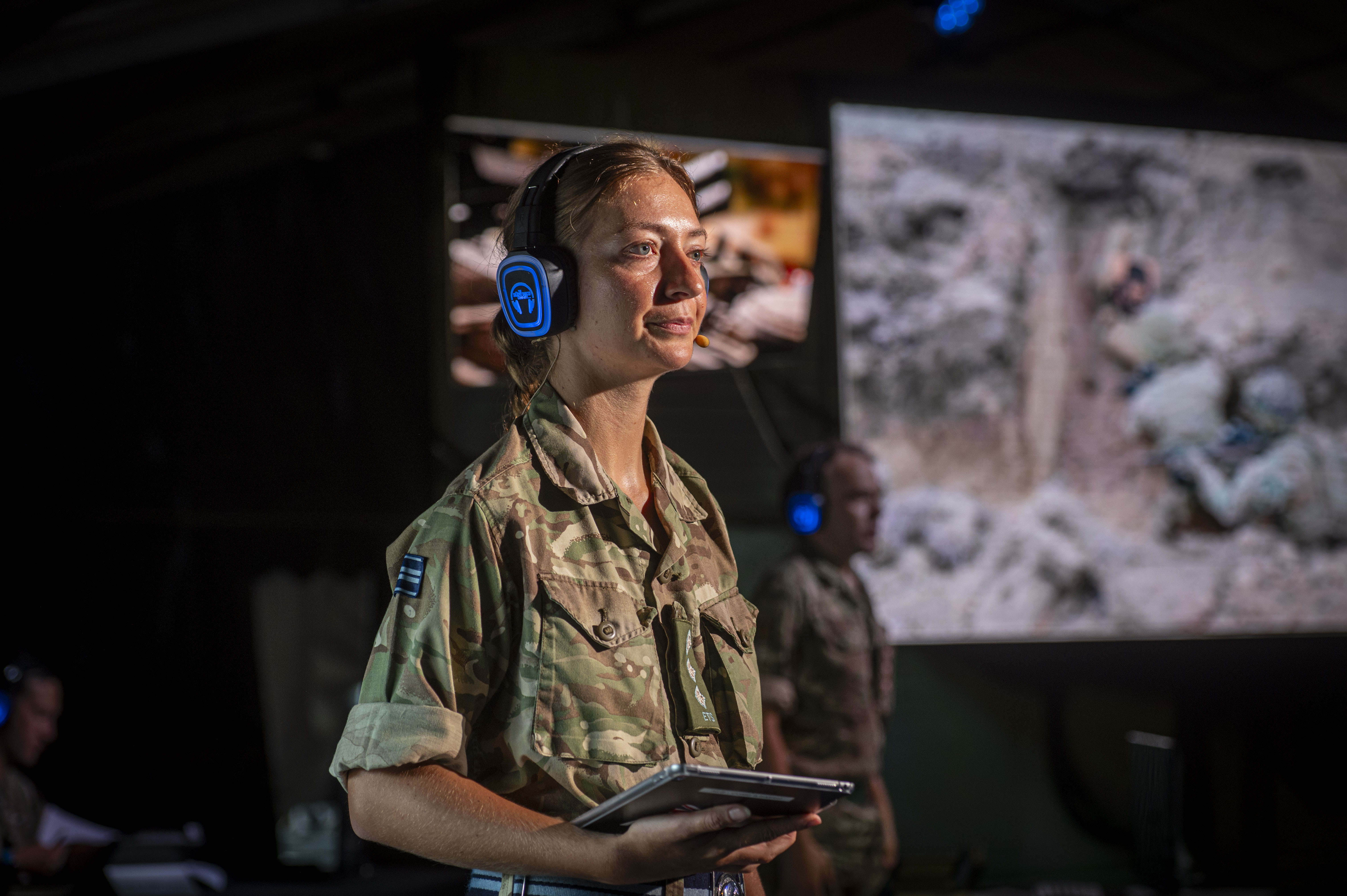 Soldier in camouflage uniform wearing blue headphones and holding a tablet during a military briefing indoors.
