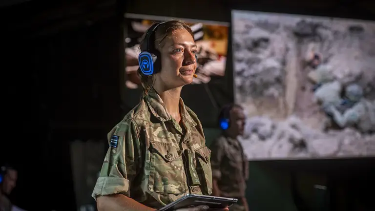 Soldier in camouflage uniform wearing blue headphones and holding a tablet during a military briefing indoors.