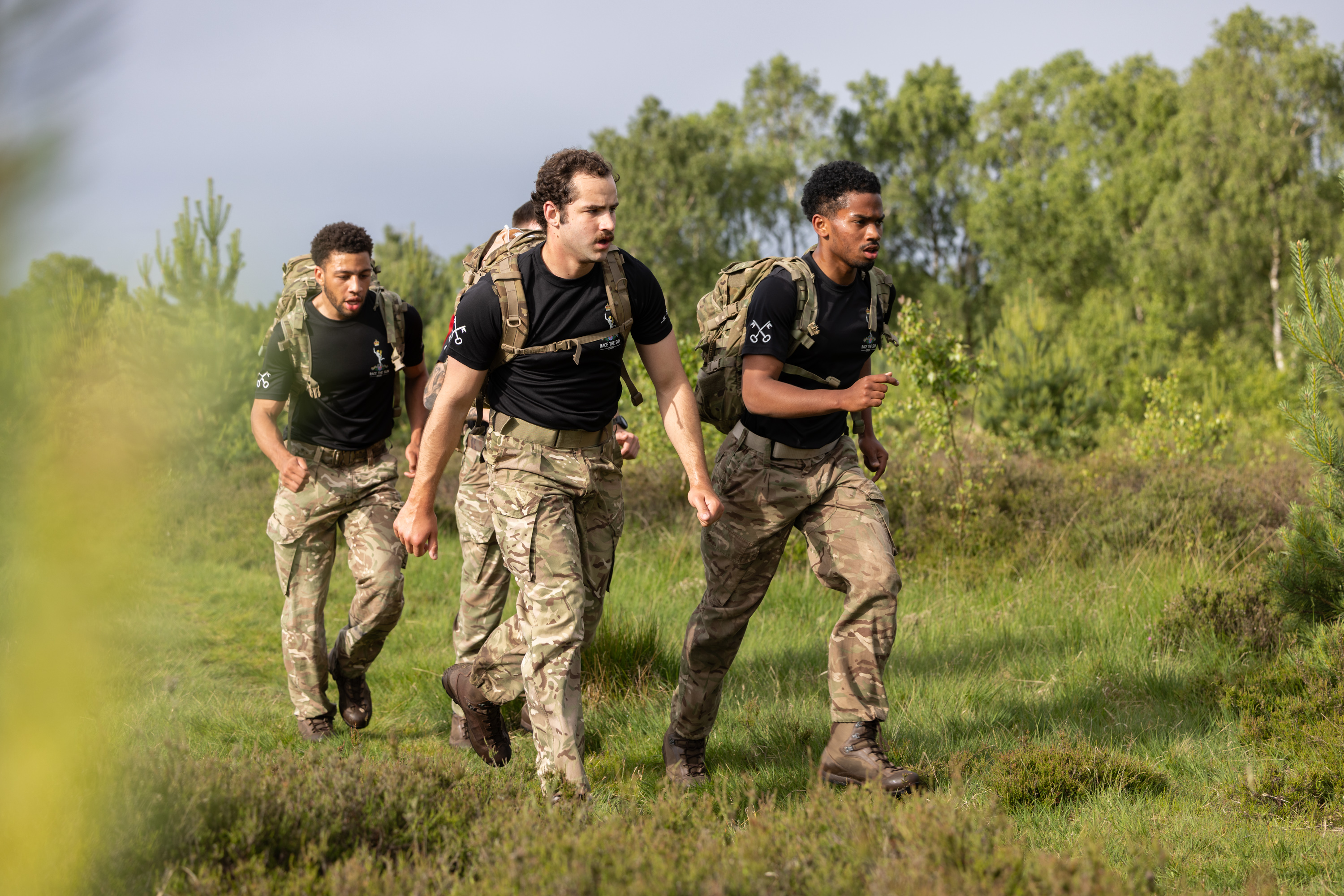 Three soldiers in camouflage uniforms and backpacks jog through a lush, green field. They convey focus and teamwork, set against a backdrop of trees.