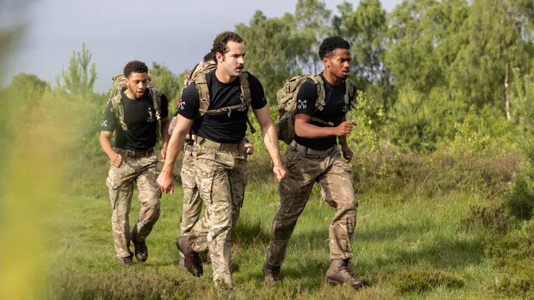 Three soldiers in camouflage uniforms and backpacks jog through a lush, green field. They convey focus and teamwork, set against a backdrop of trees.