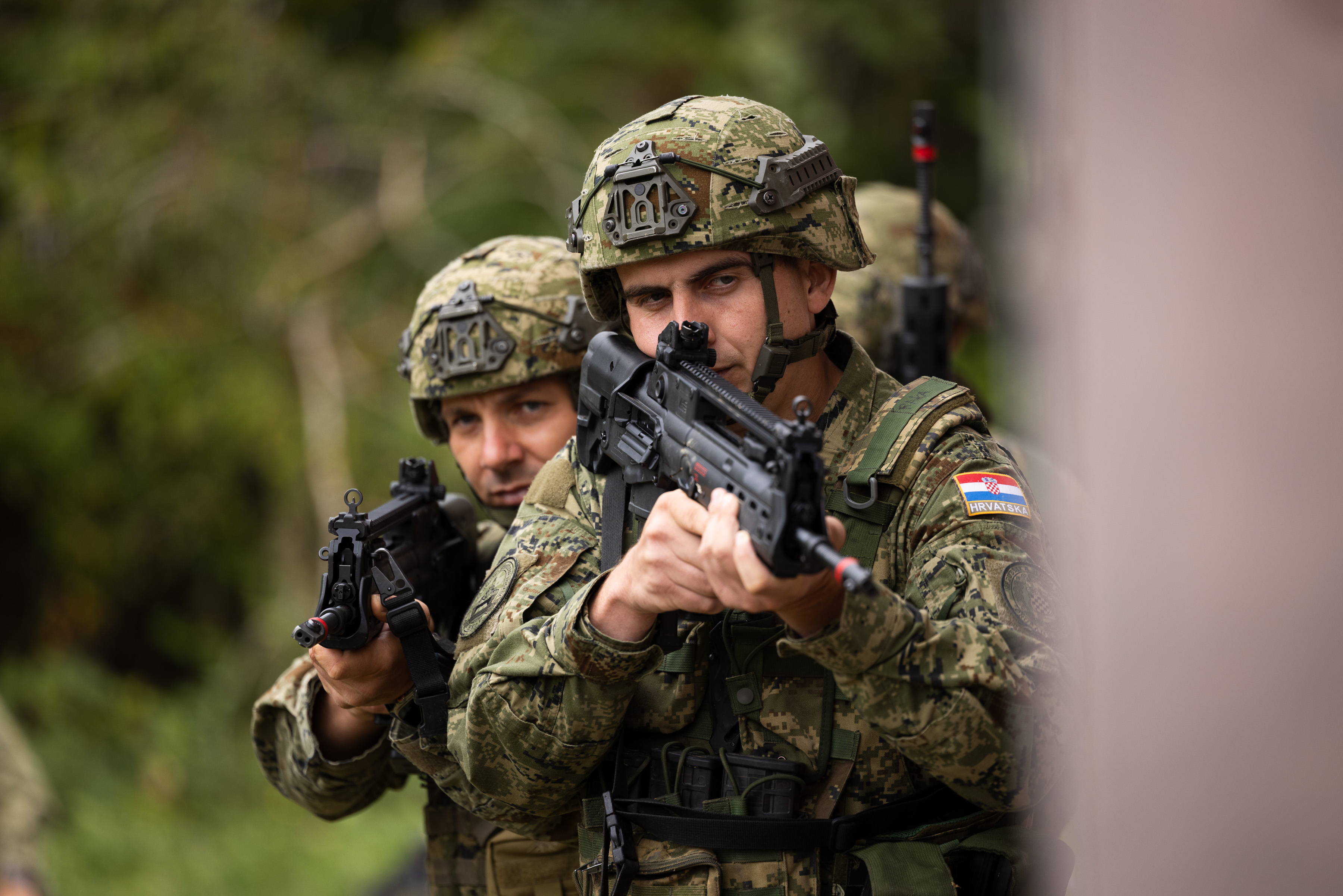 Two Croatian soldiers walking around the building in a training area wearing camouflage uniform and holding their rifles ready.