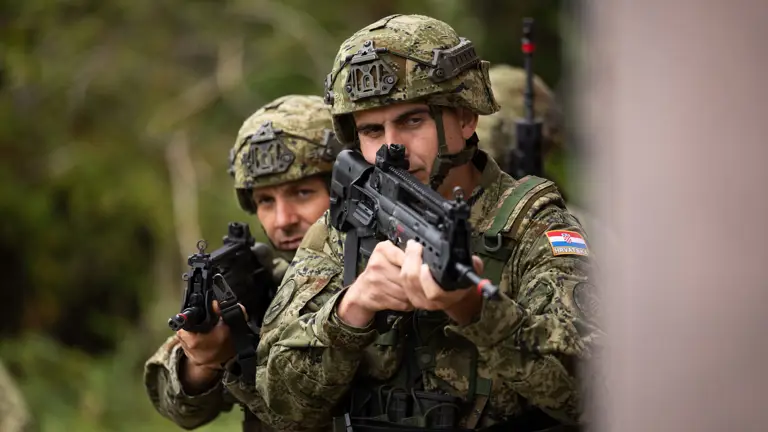 Two Croatian soldiers walking around the building in a training area wearing camouflage uniform and holding their rifles ready.