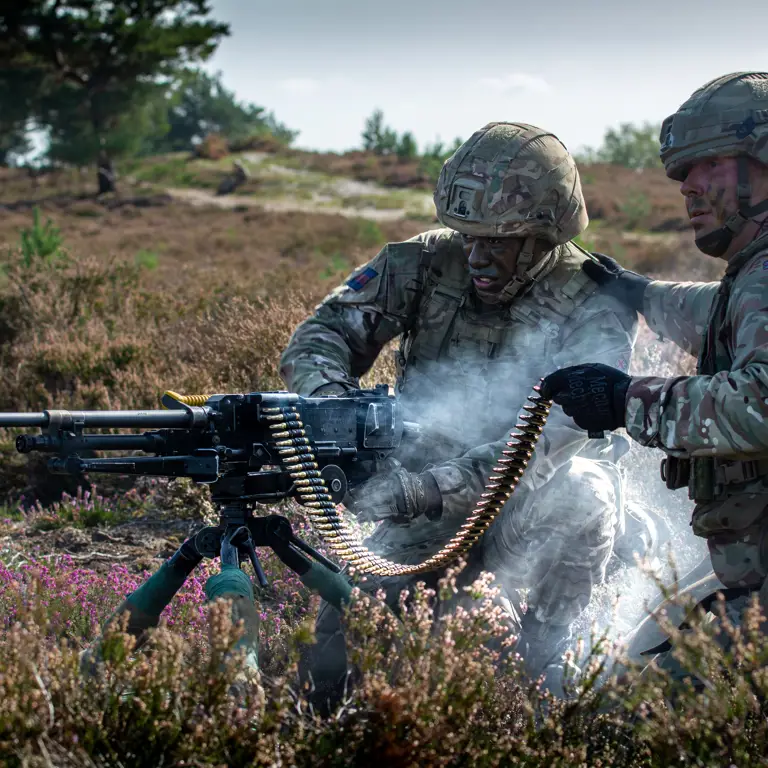 Two soldiers, wearing camouflage uniforms and helmets, kneel behind a machine gun while firing it.