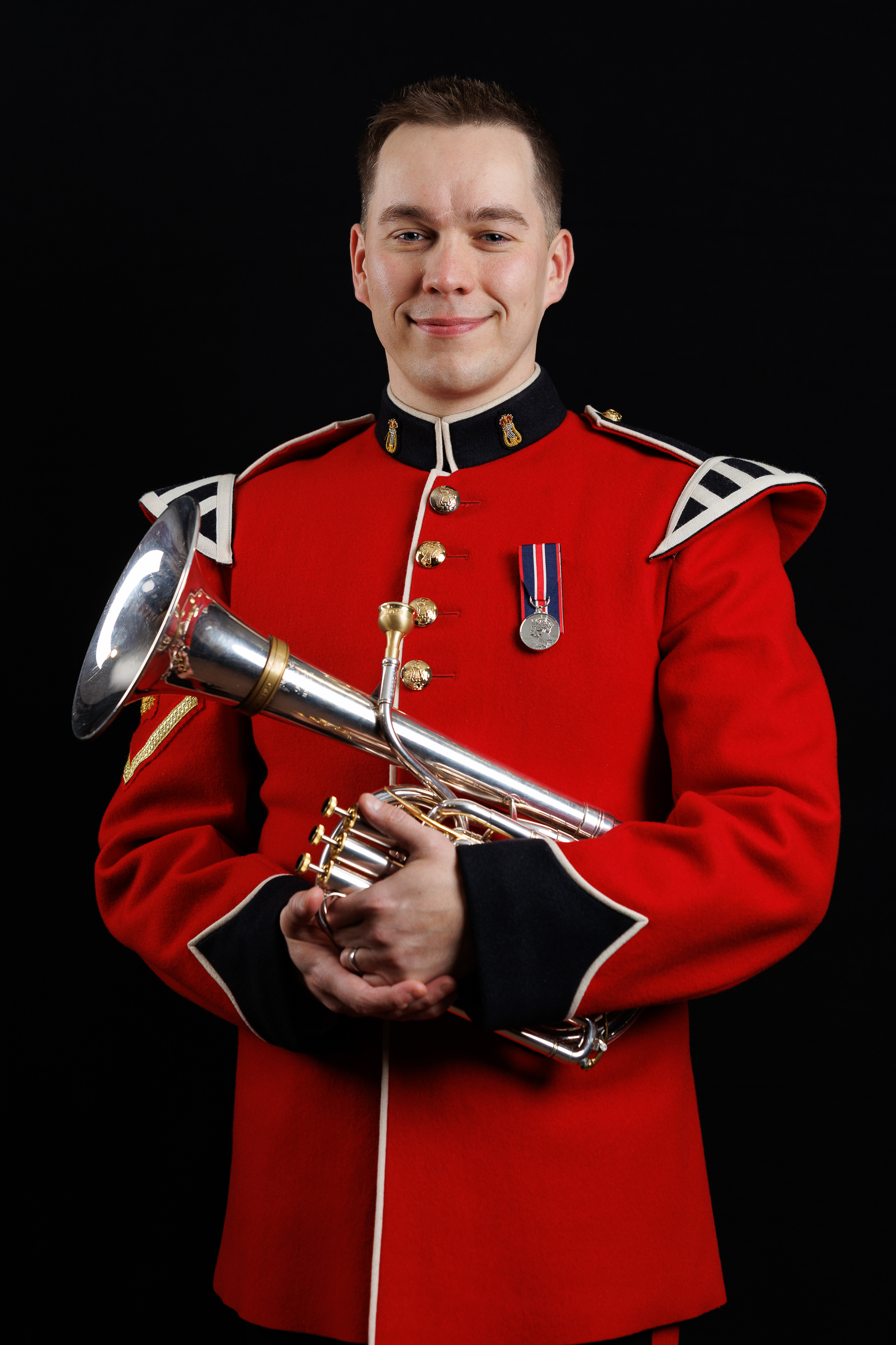 Musician in a red ceremonial military uniform holding a silver bugle against a black background.