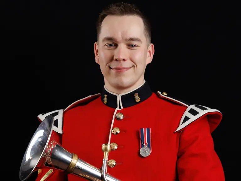 Musician in a red ceremonial military uniform holding a silver bugle against a black background.