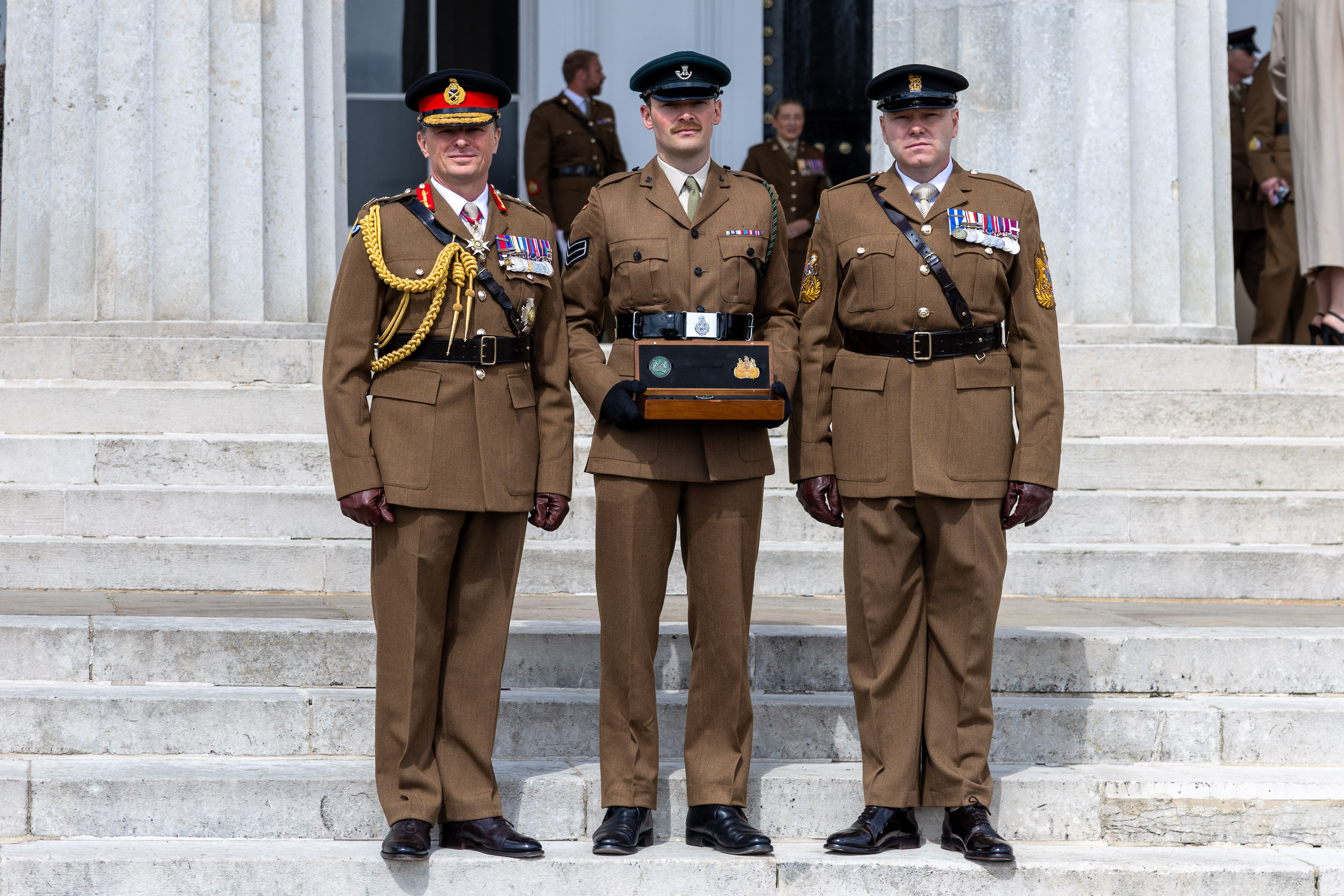 Three deocrated soliders in their hats and brown uniforms stand on the steps outside of a stone building. The middle man carried a small wooden box.