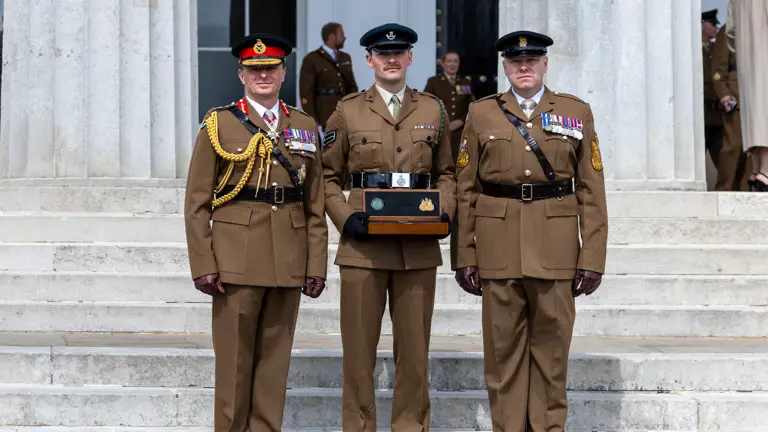 Three deocrated soliders in their hats and brown uniforms stand on the steps outside of a stone building. The middle man carried a small wooden box.