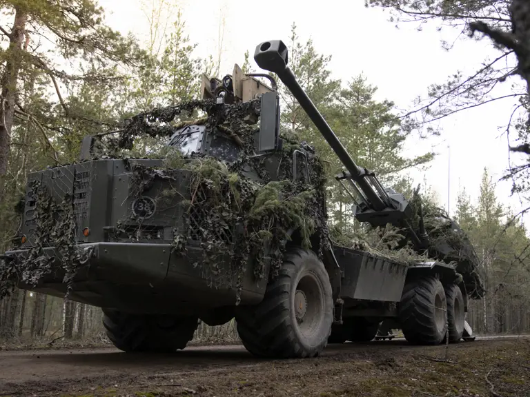 A military vehicle covered with camouflage netting and foliage is parked on a dirt road in a forested area.