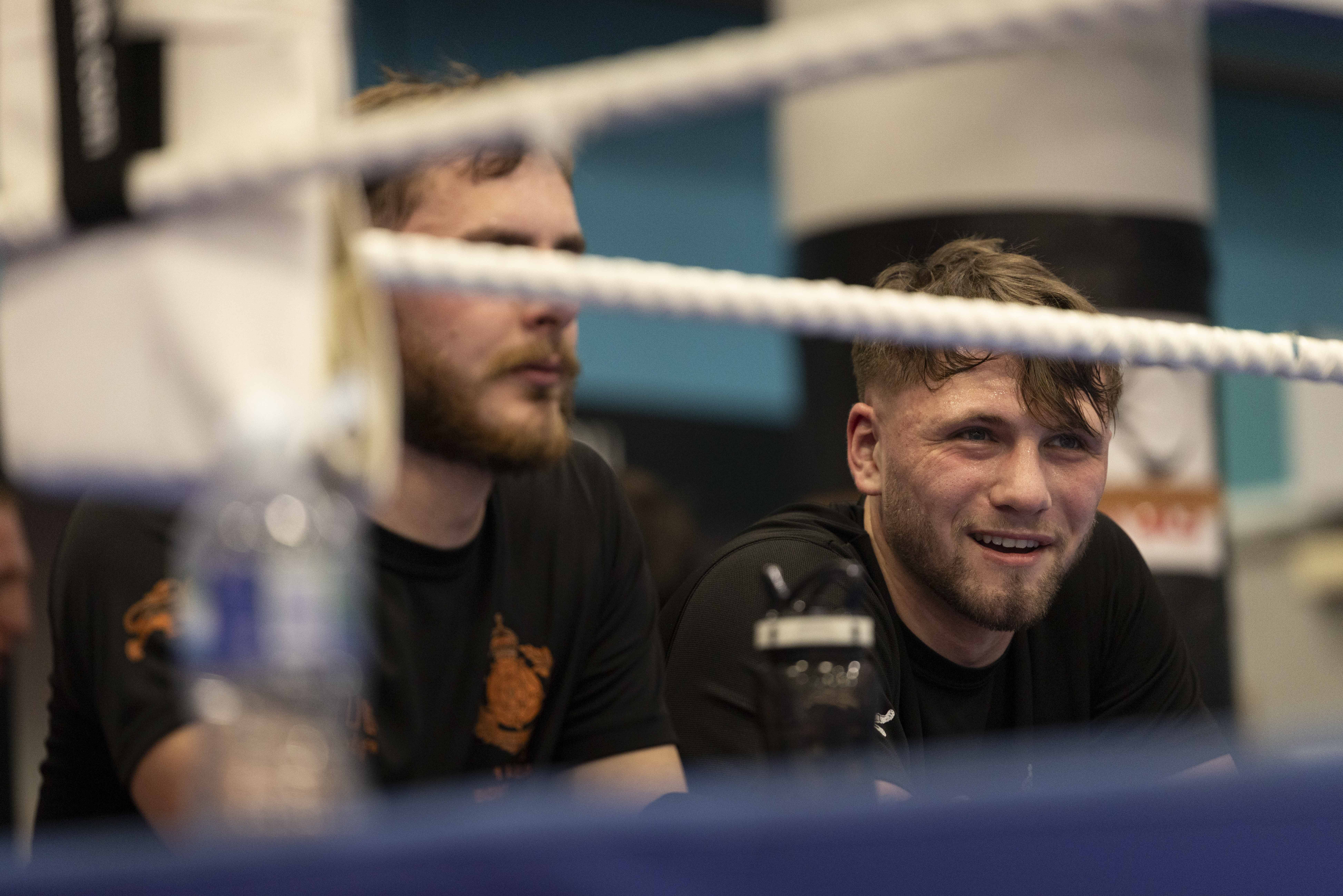 A man in a black t-shirt sits looking into a boxing ring, smiling.