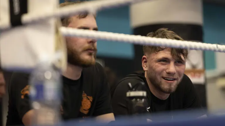 A man in a black t-shirt sits looking into a boxing ring, smiling.