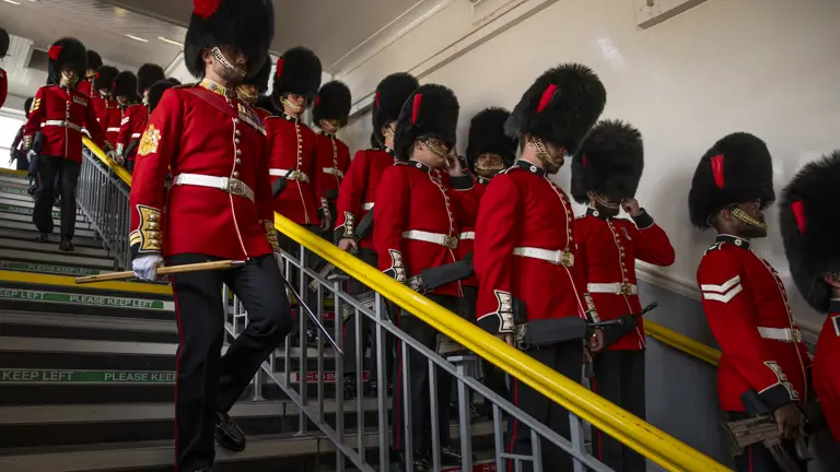 Soldiers in red tunics and black bearskin hats walk down stairs at a train station.