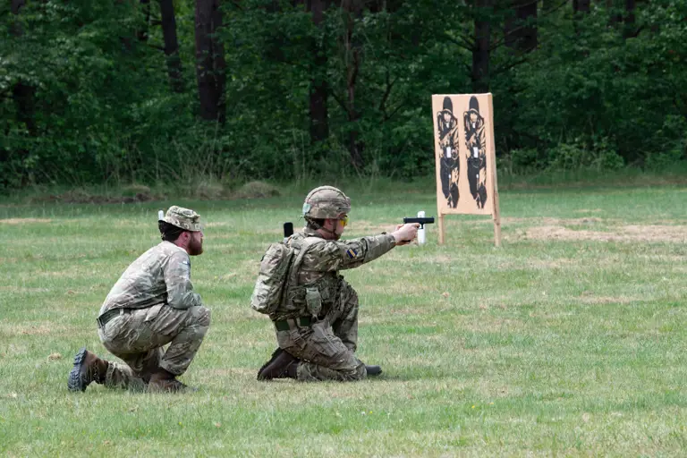 Officer Cadet on his knee in camouflage uniform firing a Pistol on the ranges under direct supervision of an instructor kneeling behind him.
