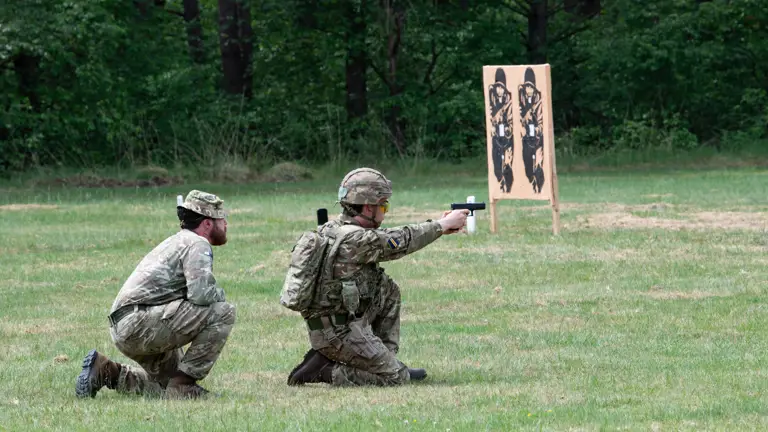 Officer Cadet on his knee in camouflage uniform firing a Pistol on the ranges under direct supervision of an instructor kneeling behind him.