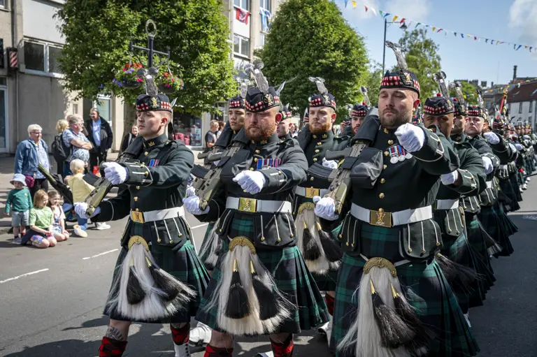 Men wearing black tunics and green and blue kilts march along a street. They are carrying rifles.