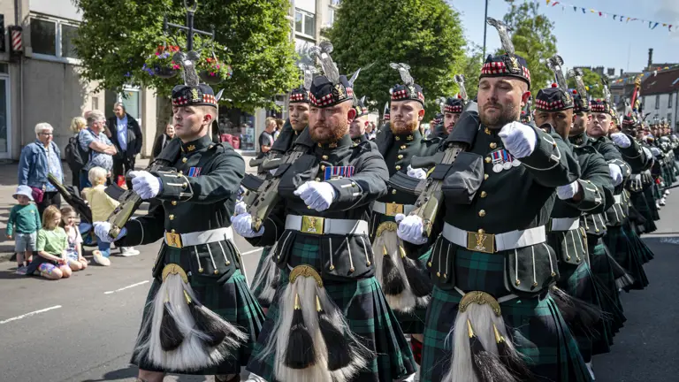 Men wearing black tunics and green and blue kilts march along a street. They are carrying rifles.