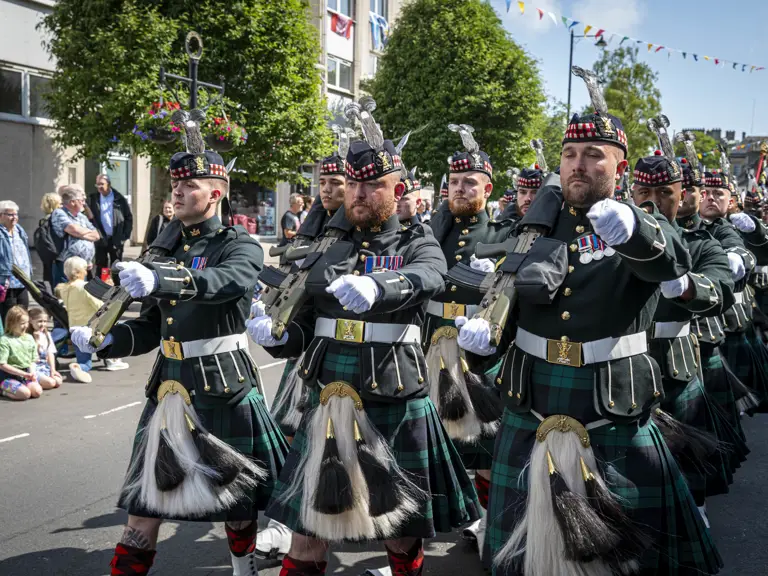 Men wearing black tunics and green and blue kilts march along a street. They are carrying rifles.