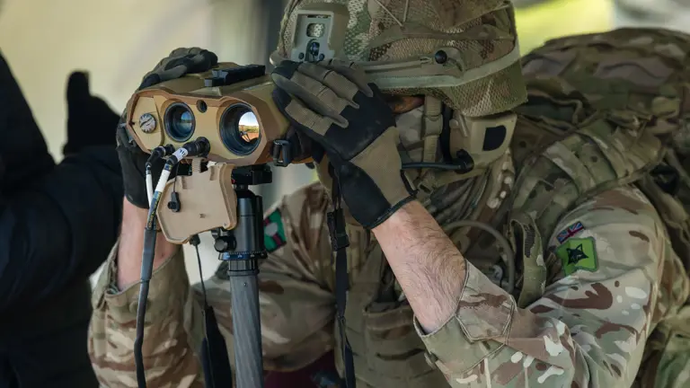 A soldier wearing camouflage uniform and a helmet looks through a set of binoculars.