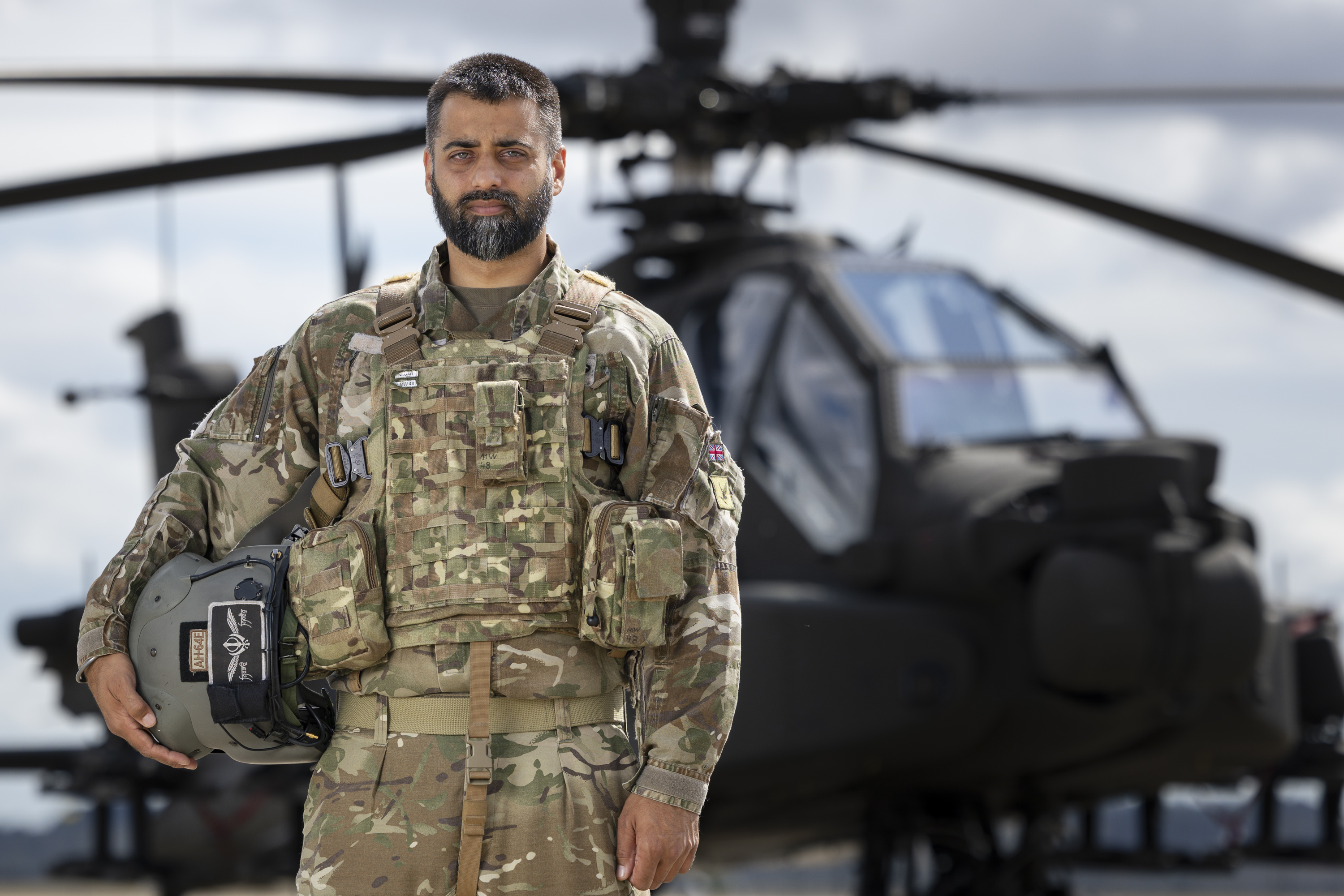 Soldier wearing camouflage uniform holding a helmet under one arm stands outside in front of an apache helicopter in the background.