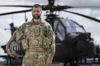 Soldier wearing camouflage uniform holding a helmet under one arm stands outside in front of an apache helicopter in the background.