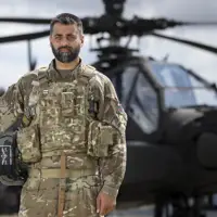 Soldier wearing camouflage uniform holding a helmet under one arm stands outside in front of an apache helicopter in the background.