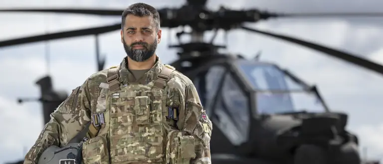 Soldier wearing camouflage uniform holding a helmet under one arm stands outside in front of an apache helicopter in the background.