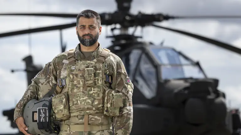 Soldier wearing camouflage uniform holding a helmet under one arm stands outside in front of an apache helicopter in the background.