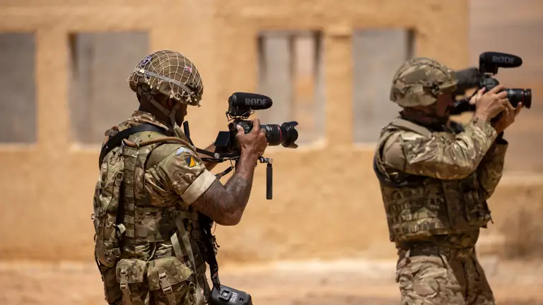 An Army Photographer stands in a sandy, arid environment filming military activity.