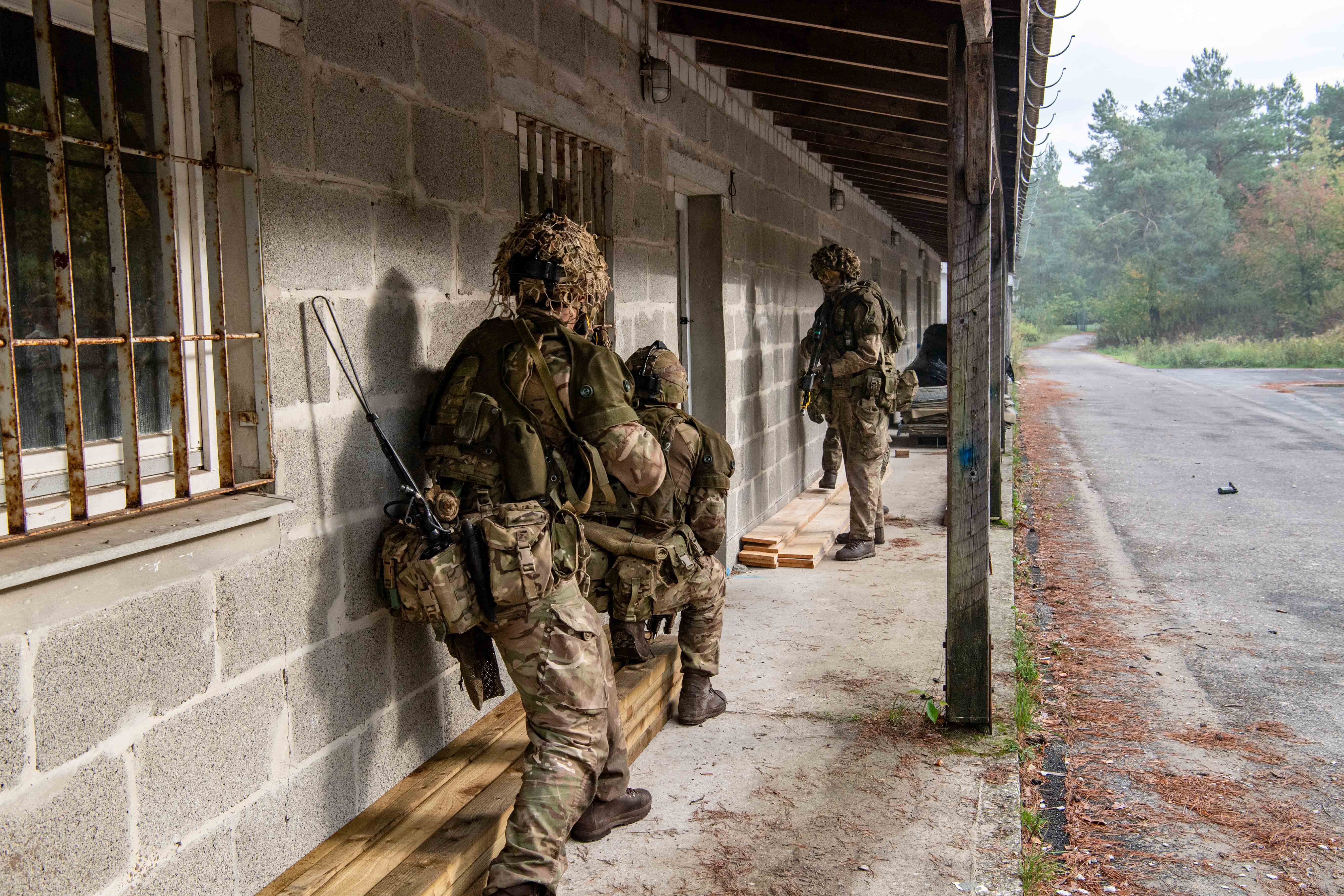 Soldiers in camouflage gear, crouched by a concrete building with barred windows, assume tactical positions.